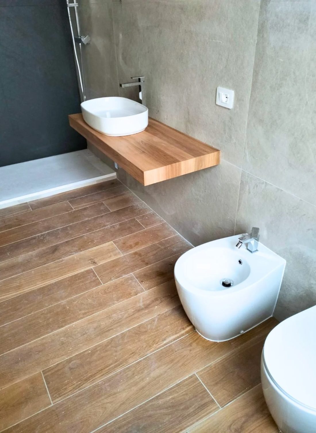 Lo stesso bagno finito, con lavabo d'appoggio su mensola in legno, bidet sospeso e doccia a filo pavimento